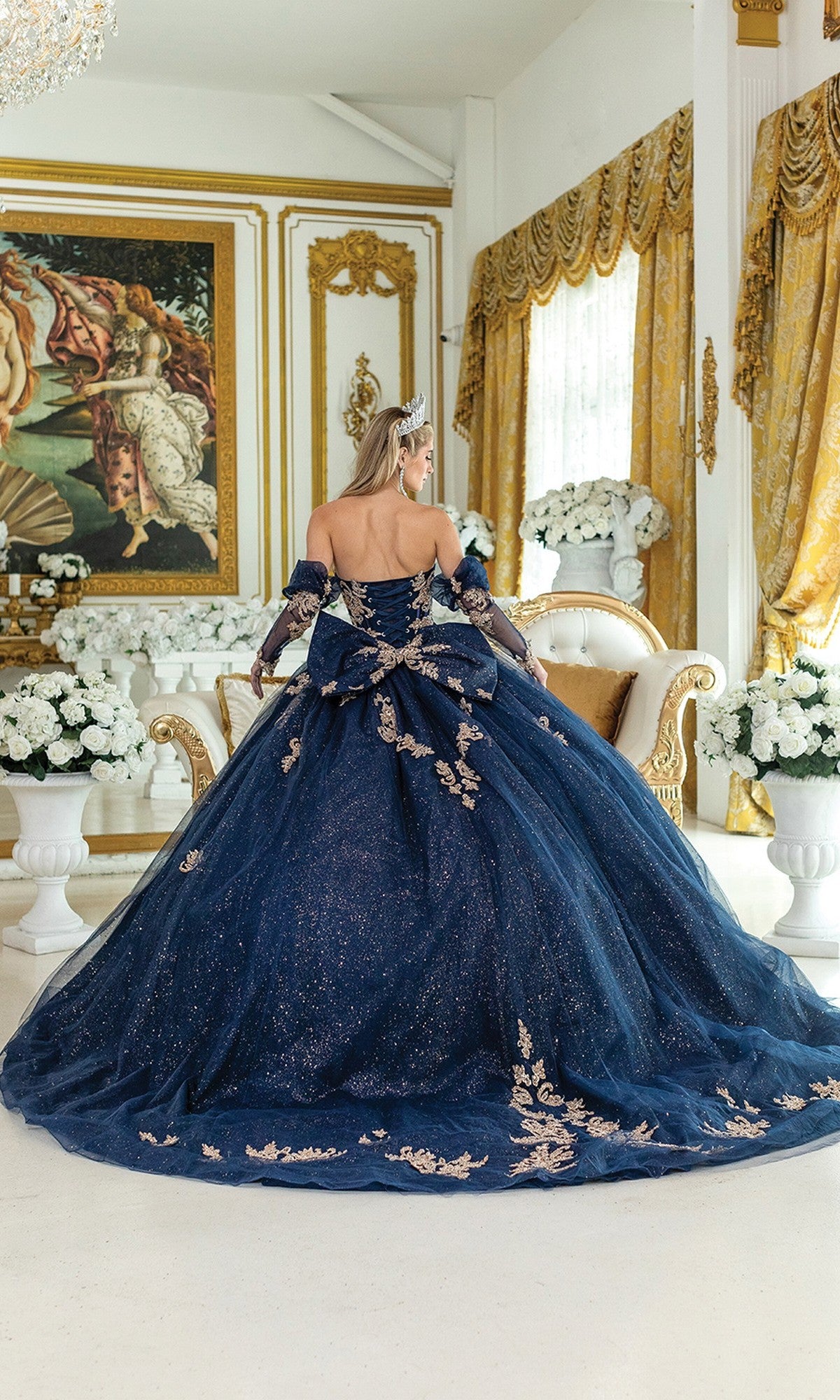 A woman in the Quinceanera Dress 5051 By Dancing Queen, a glittering navy blue tulle gown, stands with her back turned in an ornate room adorned with gold accents, white flowers, and a large classical painting on the wall.